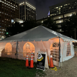 A large white event tent is set up in a city parking lot at night, with "Lot Closed" sign and traffic cones in front.