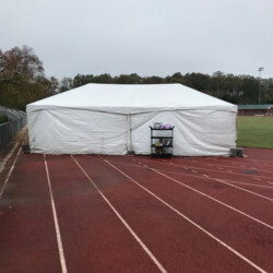 A large white tent set up on a running track, with a small rolling cart nearby, under an overcast sky.