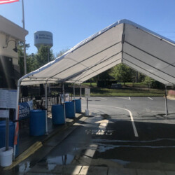 Drive-thru tent with blue barrels lined up, near a parking lot. An American flag and a water tower are in the background.