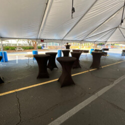 Outdoor tent with high tables covered in black cloth on an asphalt surface, set up for an event.