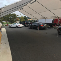 Cars lined up under a large canopy on a paved road with trees and buildings in the background.