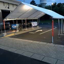 A large white tent set up over a paved area in a parking lot, with banners hanging on the side and cones marking the entrance.