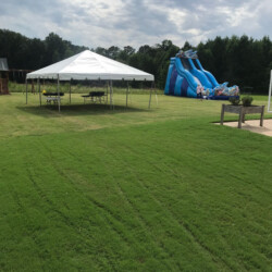 A grassy field with a white tent and picnic tables. A blue inflatable slide stands in the background under a cloudy sky.