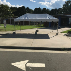 A white canopy tent is set up on a sidewalk outside a building with a cloudy sky and trees in the background.