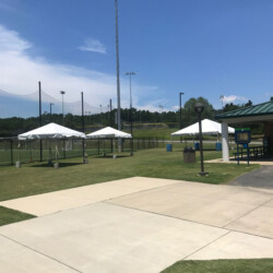 A sports field with metal fences, outdoor lights, white tents, and a low building under a clear blue sky.