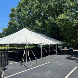 A large white tent is set up in a parking lot near trees on a clear day, with tables and support poles visible underneath.