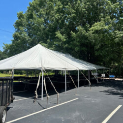 A large white tent is set up in a parking lot near trees on a clear day, with tables and support poles visible underneath.