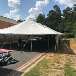 Large white tent set up outdoors on pavement near a building, with trees and cloudy sky in the background.