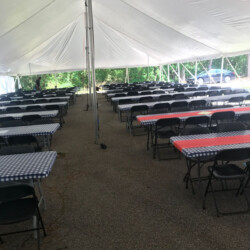 Large outdoor tent with rows of tables covered in checkered tablecloths, accompanied by folding chairs.