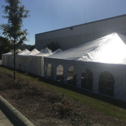 Row of white tents set up outside a building, with trees and a parking lot visible under a clear blue sky.