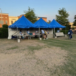 Three blue tents set up outdoors with people seated underneath, positioned on a grassy area near brick buildings and trees.
