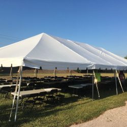 A large white tent set up outdoors over rows of folding tables and chairs on grassy ground, under a clear blue sky.
