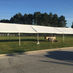 A large white tent is set up on a grassy area near a paved surface, with trees in the background under a clear blue sky.