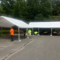 Workers set up white tents in a parking lot, preparing tables underneath, surrounded by trees.