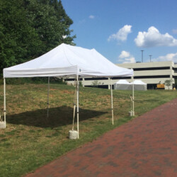 A white canopy tent is set up on a grassy area near a red brick path and a parking structure under a blue sky.