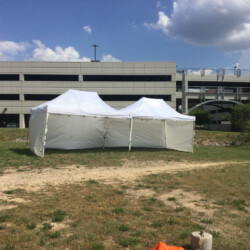 Two white pop-up tents set up on a grassy area near a multi-story parking garage under a partly cloudy sky.