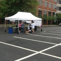 People gather under a white tent in a parking lot near a brick building, with tables and a banner visible.