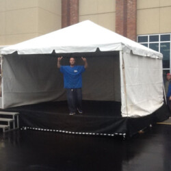 Person standing and posing inside a white tent with a small stage, outdoors near a building with brick walls.