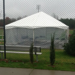 A large white tent is set up on a grassy field, surrounded by fencing. Benches are in the foreground, and soccer goals are visible.
