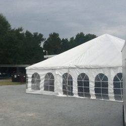 A large white tent with arched windows stands on a gravelly surface, surrounded by trees under a cloudy sky.