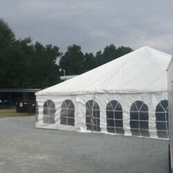 A large white tent with arched windows stands on a gravelly surface, surrounded by trees under a cloudy sky.