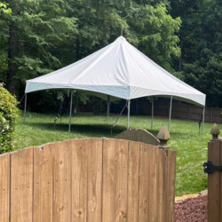 A white outdoor tent is set up on a grassy lawn, surrounded by trees and a wooden fence.