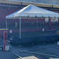 A white canopy tent covers a small stage on a concrete surface, with surrounding equipment and a brick building in the background.