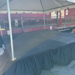 Outdoor stage with a black skirt and overhead canopy, set up on a paved area with a building and benches in the background.