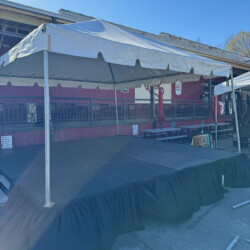 Outdoor stage with a canopy and black skirt near a building, set up on a paved area with some tables in the background.
