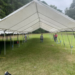 Several people are setting up a large outdoor tent in a grassy area with surrounding trees.