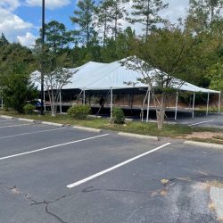 Large white event tent set up in an empty parking lot surrounded by trees under a partly cloudy sky.