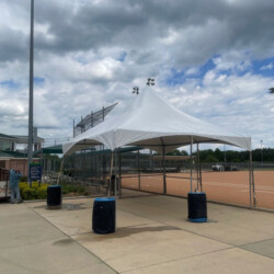 A white canopy tent stands on a paved area near a chain-link fence under a cloudy sky, with four sandbag weights at the corners.