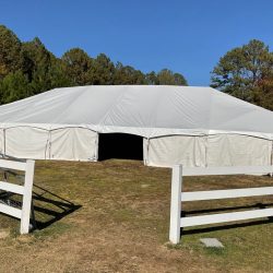 A large white tent is set up on grass, surrounded by a white wooden fence, with trees and a clear blue sky in the background.