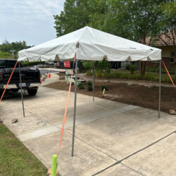 A white canopy tent set up on a concrete sidewalk near a parked black truck, with trees and a lawn in the background.