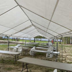 Empty outdoor tent with folding tables and chairs set up on a grassy surface, surrounded by trees and an overcast sky.
