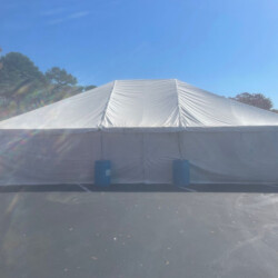 Large white tent with a pointed roof, secured by blue barrels, set up on a paved area under a clear blue sky.