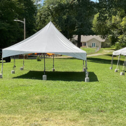 A white tent on a grassy lawn, secured with concrete blocks, with trees and a house in the background.