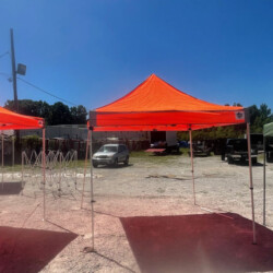 An orange canopy tent set up outdoors on a gravel surface, with cars and trees in the background under a clear blue sky.