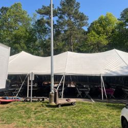 Large white event tent set up outdoors on grass, with tables and chairs visible underneath, surrounded by trees and a clear sky.