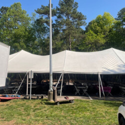 Large white event tent set up outdoors on grass, with tables and chairs visible underneath, surrounded by trees and a clear sky.