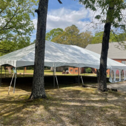 A large white event tent is set up outdoors between trees, with a red brick building visible in the background.