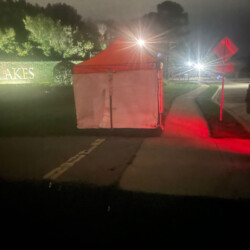 A small white tent with an orange top on a sidewalk at night, illuminated by nearby streetlights and car lights.