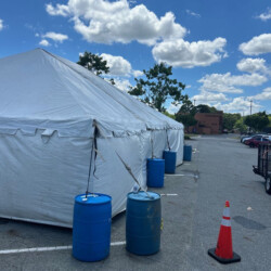 A large white tent secured with blue barrels is set up in a parking lot on a clear day, with an orange cone nearby.