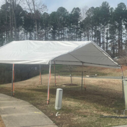 A white canopy tent set up outdoors on grass near a sidewalk, with tall trees in the background.