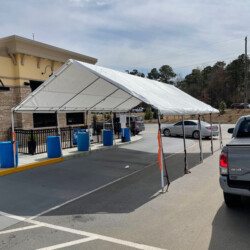 White canopy tent set up in a parking lot with blue trash bins and cars nearby on a cloudy day.